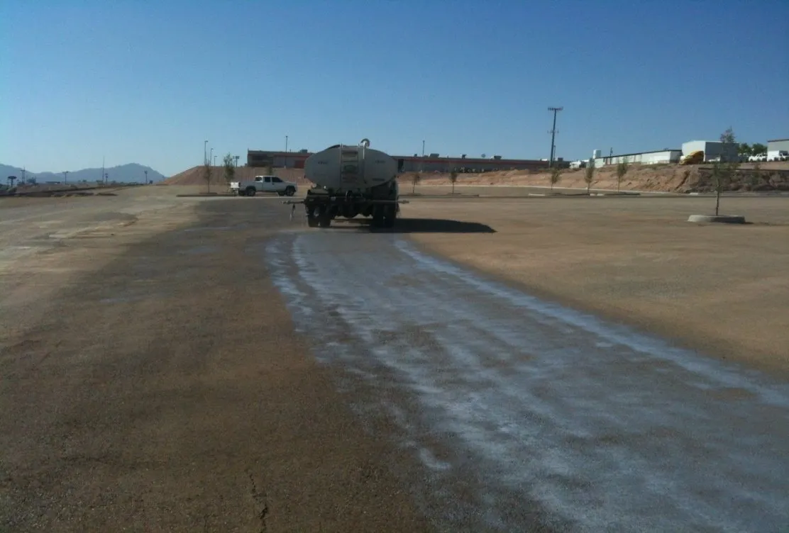 A water truck spraying diluted product across a wide unpaved lot, with a pale wet stripe behind it