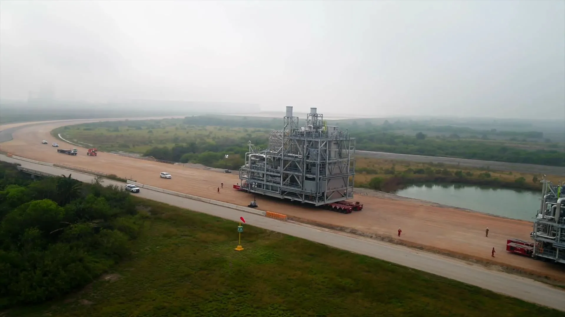 Two large industrial process modules on multi-axle transporters, moving along a wide unpaved access road beside a pond