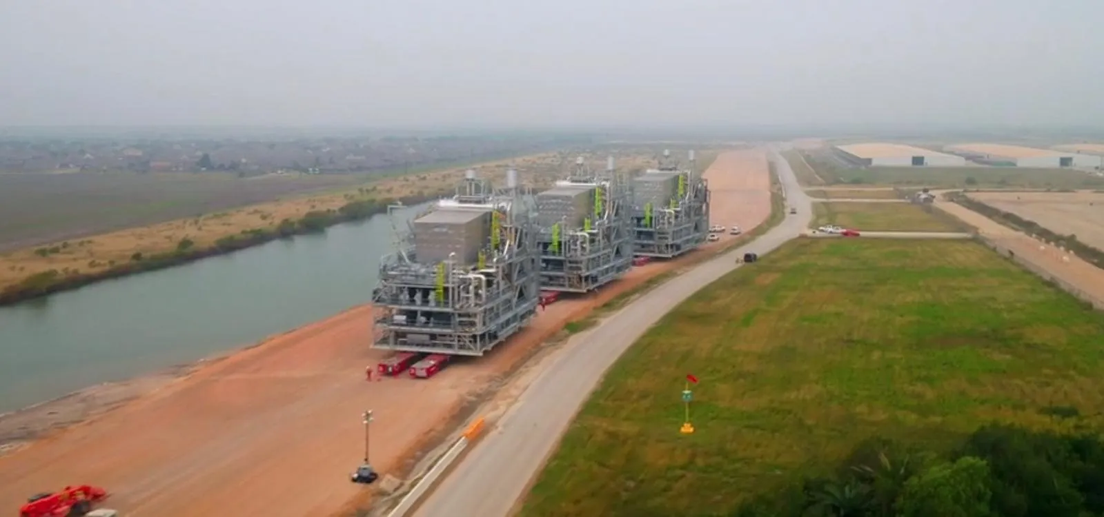Three large industrial process modules on transporters, moving along a wide unpaved access road beside a canal