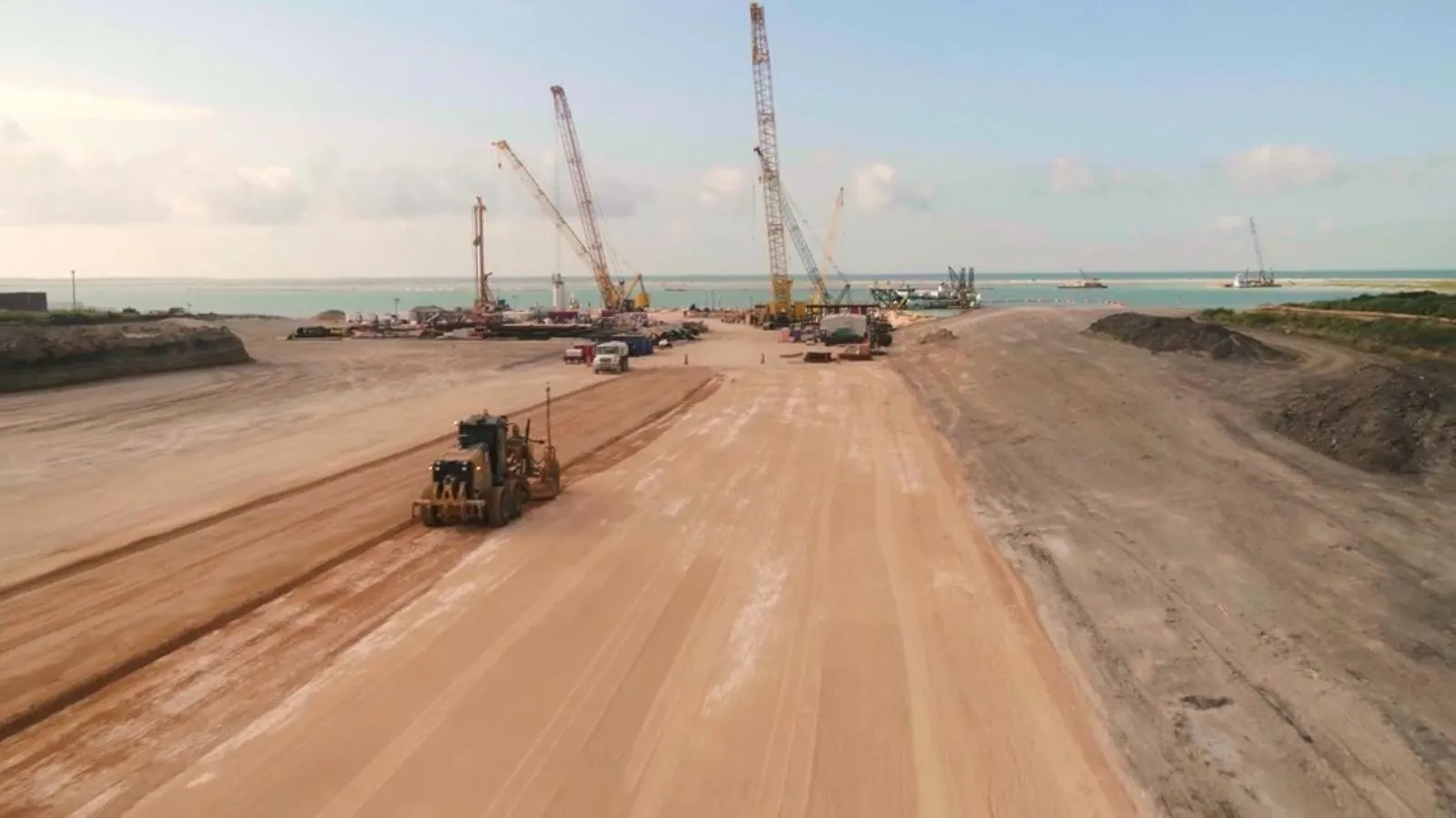 A motor grader working a wide unpaved road base on an industrial site, with cranes along the shoreline behind