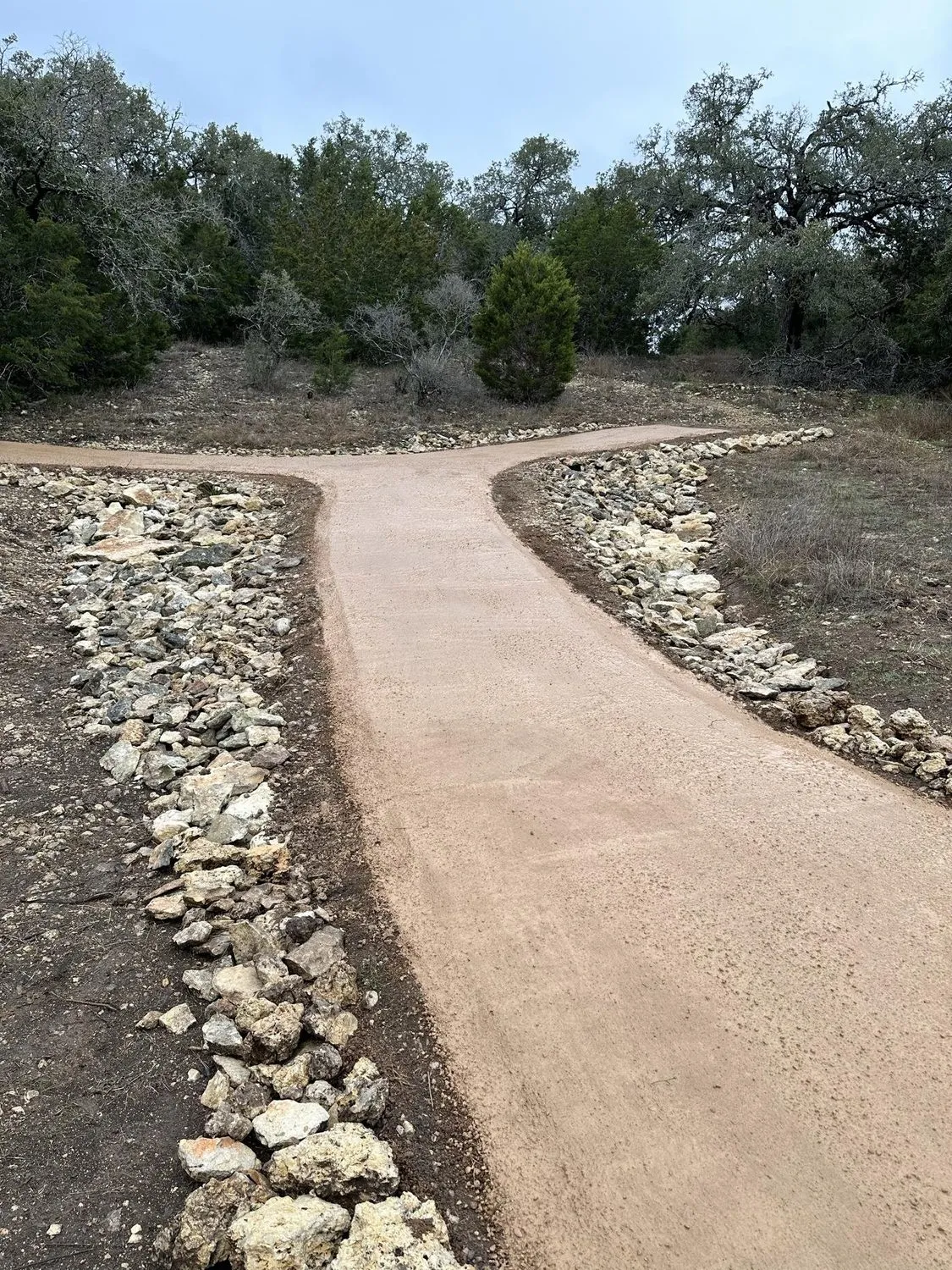 A finished, smooth unpaved private road edged with limestone rock, branching through oak and cedar trees